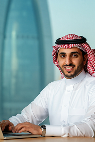 A smiling Arab businessman wearing a white thobe and red checkered shemagh, sitting at a desk with a laptop in a modern office setting, with a glass skyscraper visible in the background.